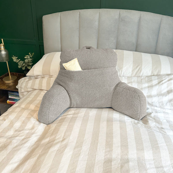 Oatmeal teddy fleece reading cushion with a large pocket with a hot water bottle in the pocket (not included), reading cushion on a bed with natural and white striped bedding (not included)