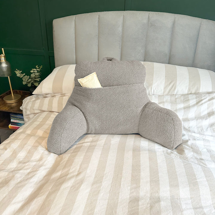 Oatmeal teddy fleece reading cushion with a large pocket with a hot water bottle in the pocket (not included), reading cushion on a bed with natural and white striped bedding (not included)